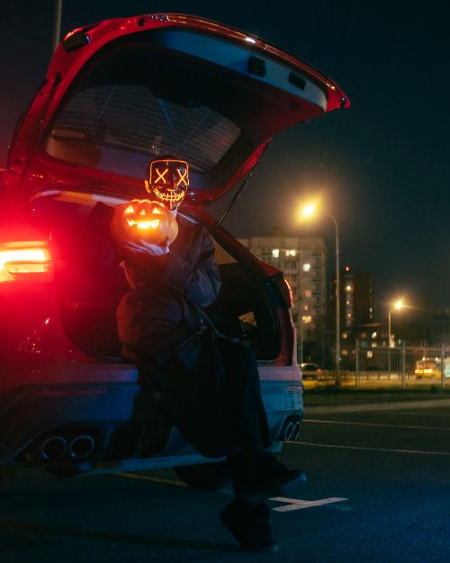 Free Masked figure holding a lit pumpkin in a car trunk, night scene with urban background. Stock Photo