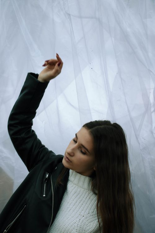 Free Young woman in black leather jacket posing with closed eyes against a soft white curtain background. Stock Photo