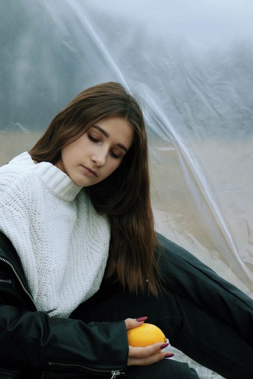 Free Young brunette woman in cozy outfit holding a lemon with a reflective expression. Plastic backdrop sets an environmental tone. Stock Photo