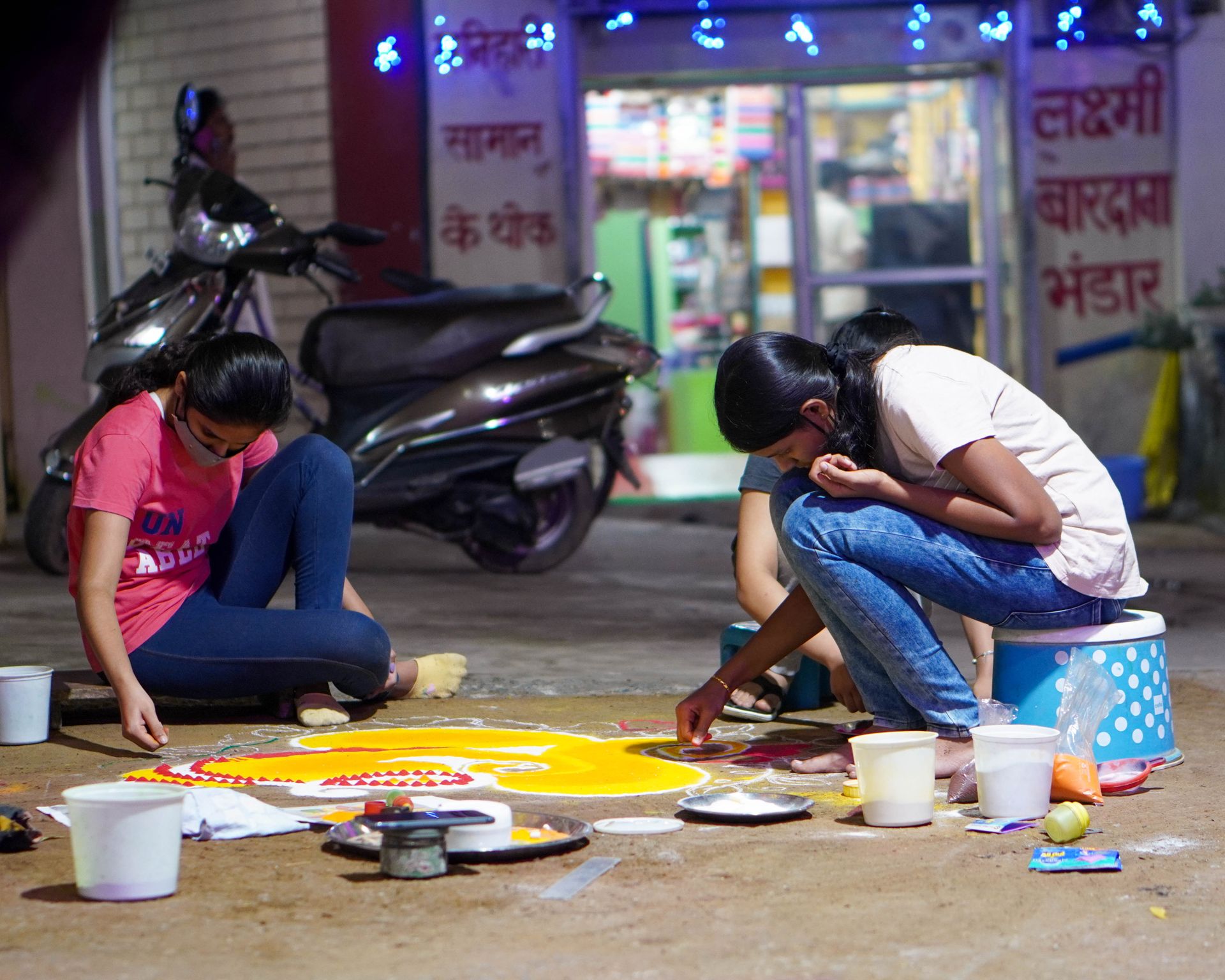 Two teenagers crafting colorful rangoli art on an Indian street during a festive night.