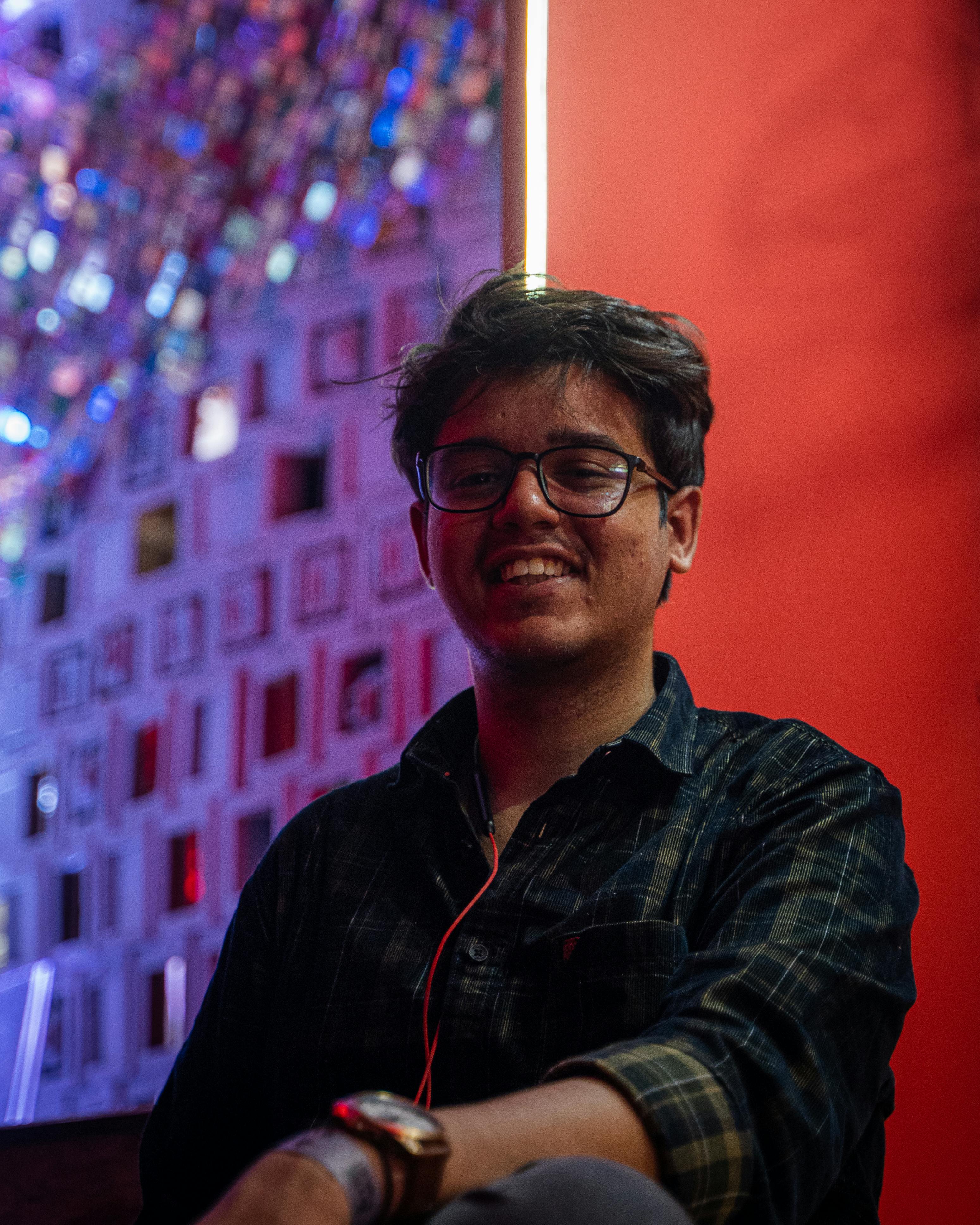 Portrait of a young man wearing glasses and a plaid shirt, smiling indoors with colorful background lighting.