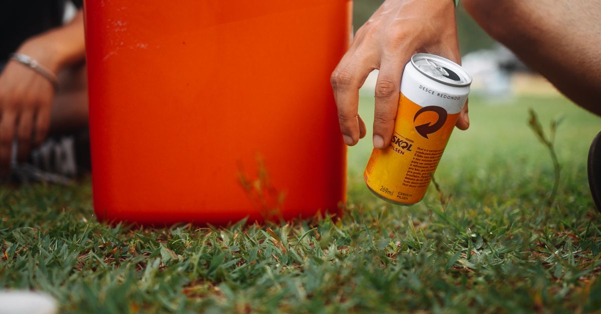 A hand holding a beverage can next to an orange cooler on green grass, signifying summer relaxation.