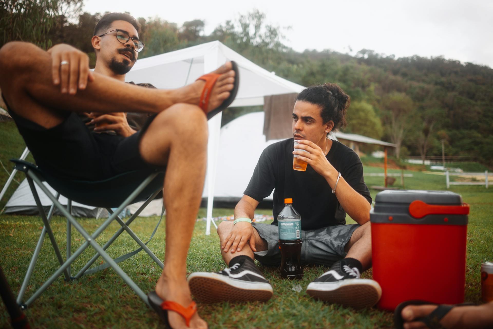 Two young men enjoying a picnic outdoors, sitting on the grass with a cooler and drinks.