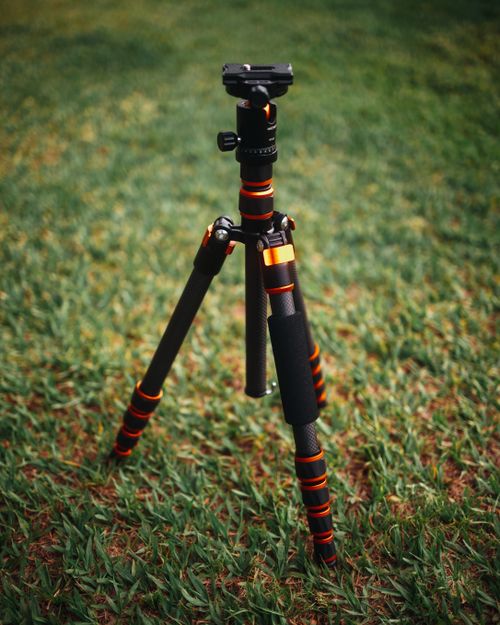 Free A sturdy black and orange carbon fiber tripod stands tall on lush green grass. Stock Photo