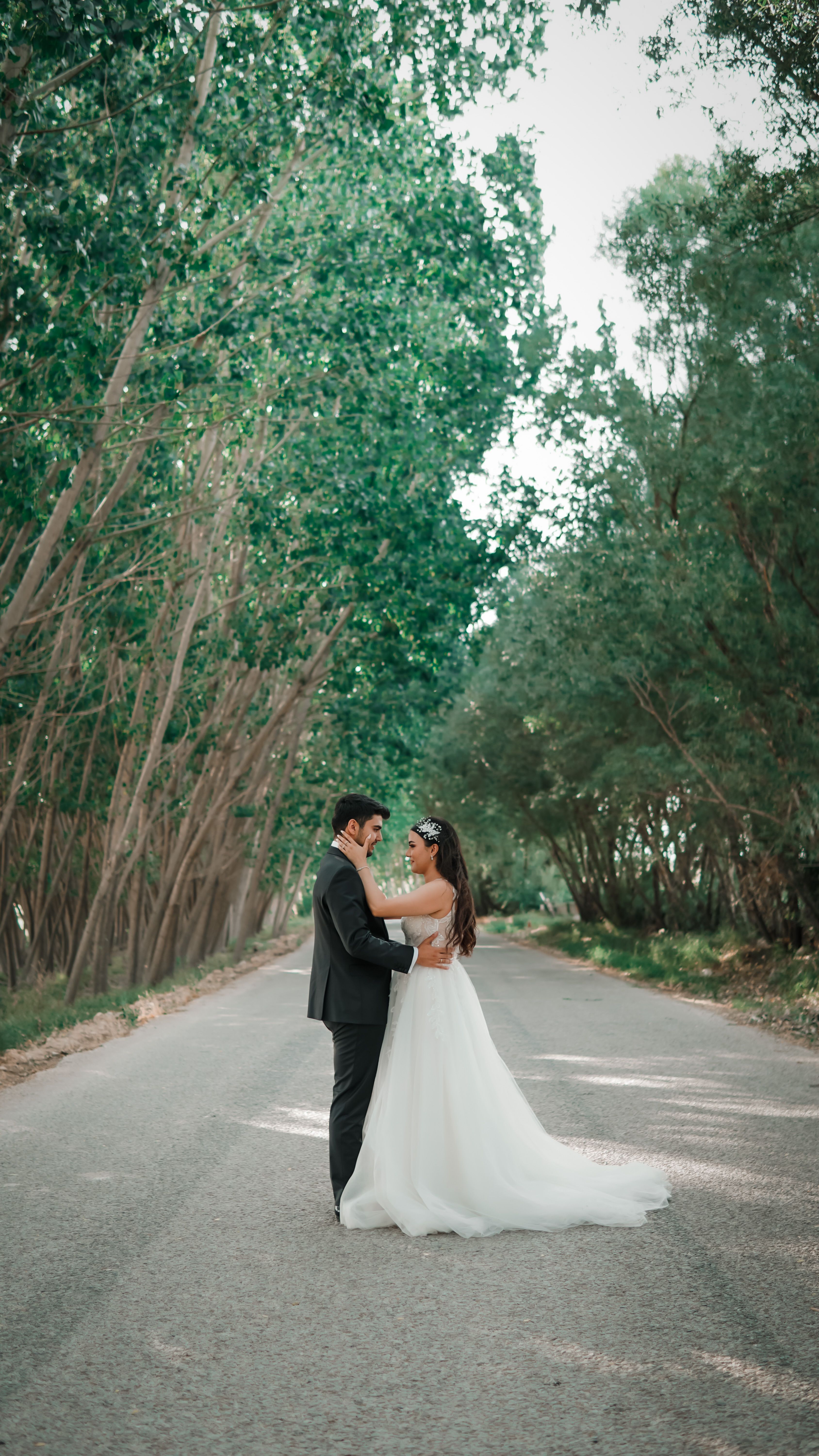 A newlywed couple embraces on a scenic tree-lined road, capturing a romantic moment.