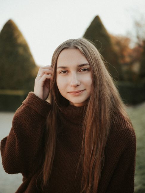 A young woman with long brown hair smiling outdoors in a garden setting at sunset.