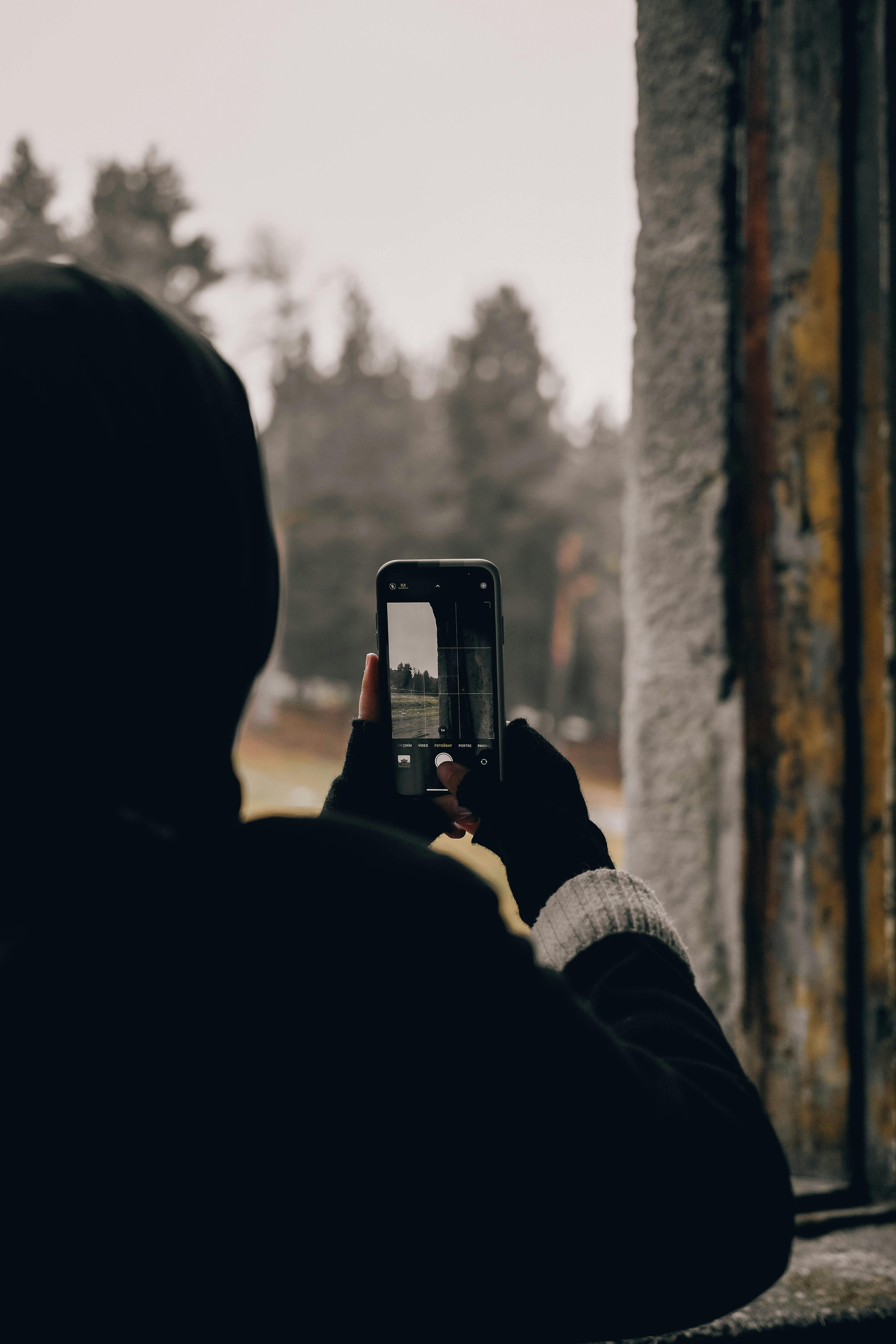Free Hooded person taking a photo of a forest scene using a smartphone through a window. Stock Photo
