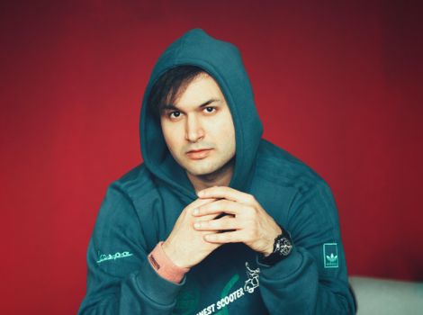 Close-up portrait of a man wearing a blue hoodie, hands together, against a vivid red background.