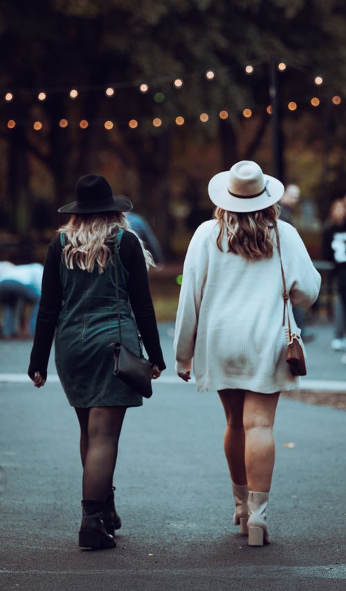 Free Two women in stylish outfits walking in a park setting, showcasing fashion accessories like hats and sling bags. Stock Photo