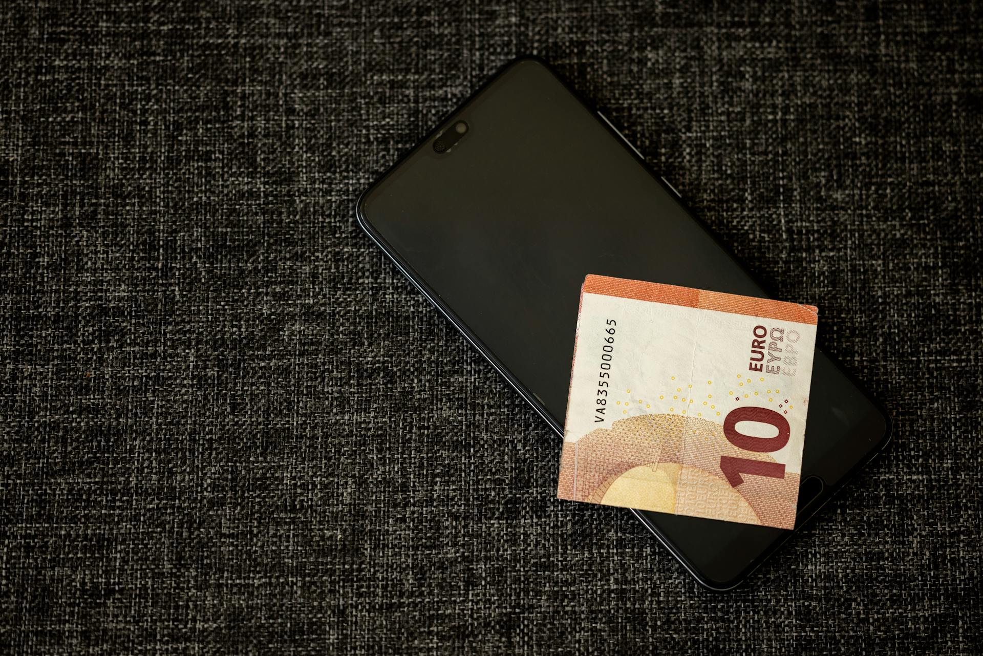 A 10 euro banknote placed on a smartphone on a textured dark fabric background.