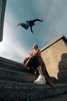 A parkour athlete leaps over a sitting woman on concrete steps under a clear sky.