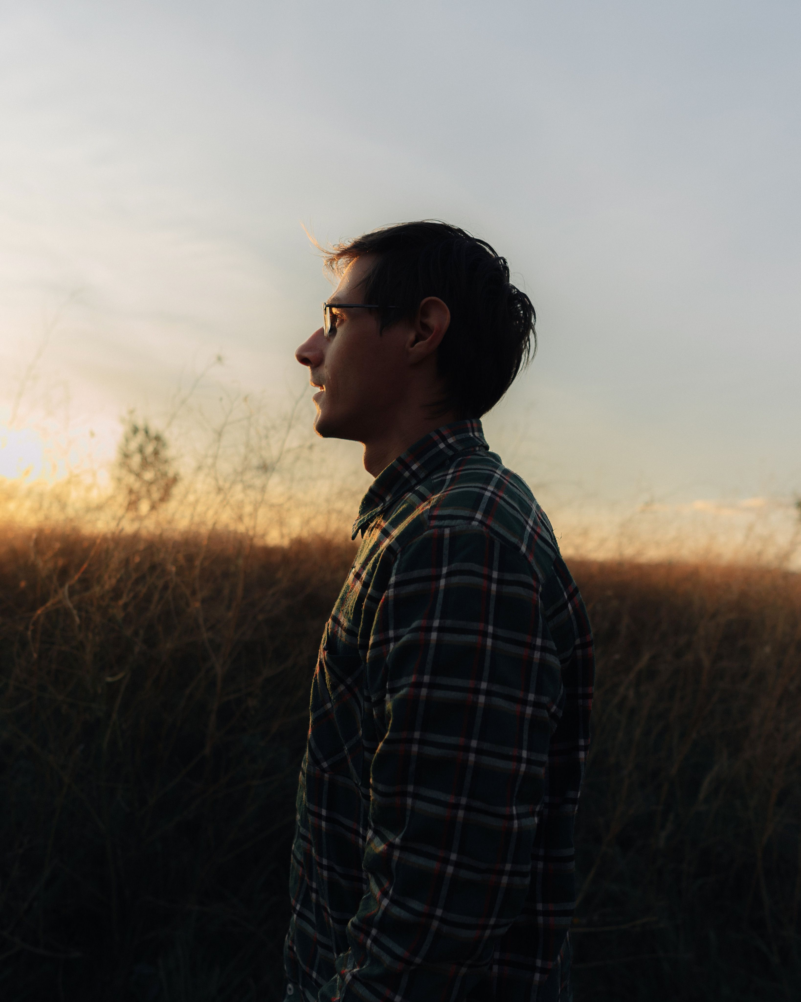 Silhouette of a man in a checkered shirt standing outdoors at sunset, captured in side profile.