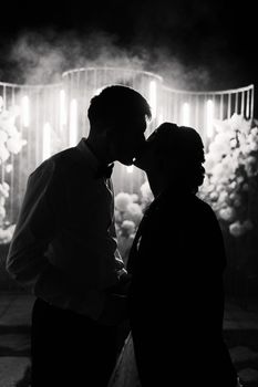 Silhouette of a bride and groom sharing a kiss in backlit wedding portrait with atmospheric night lighting.