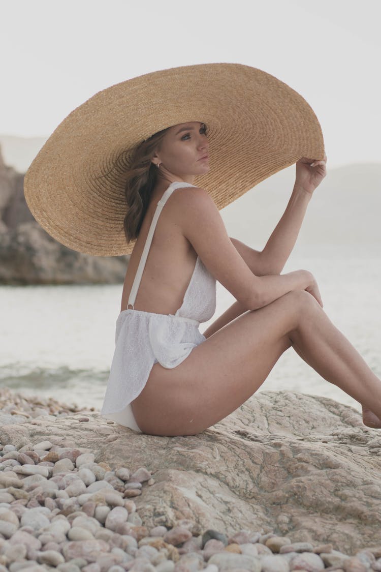 A Female Sitting On Rock Wearing A Big Hat And Beach Clothes