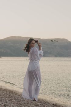 Serene scene of a woman in white dress strolling along the beach at sunset with seagulls flying.