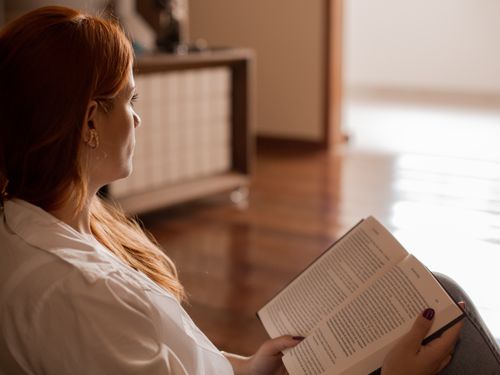 Free A woman with red hair reading a book while seated indoors on a wooden floor. Stock Photo