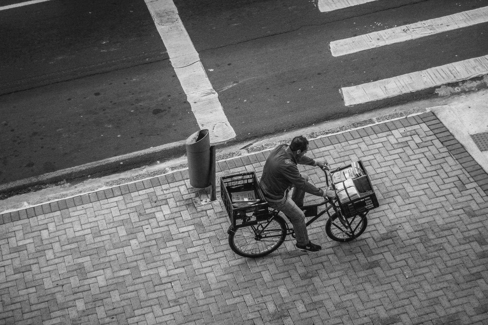 A man rides a cargo bicycle on a city sidewalk, captured from above in black and white.