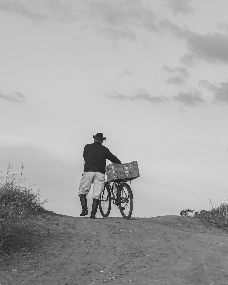 Senior Man In Hat Wheeling His Bike Uphill On Dirt Road