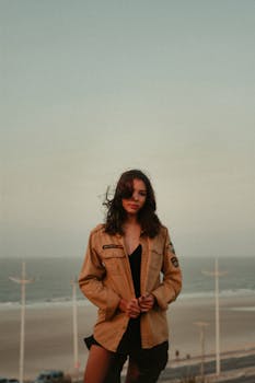 Stylish woman in a jacket stands on a beach at sunset, with a windy ambiance.