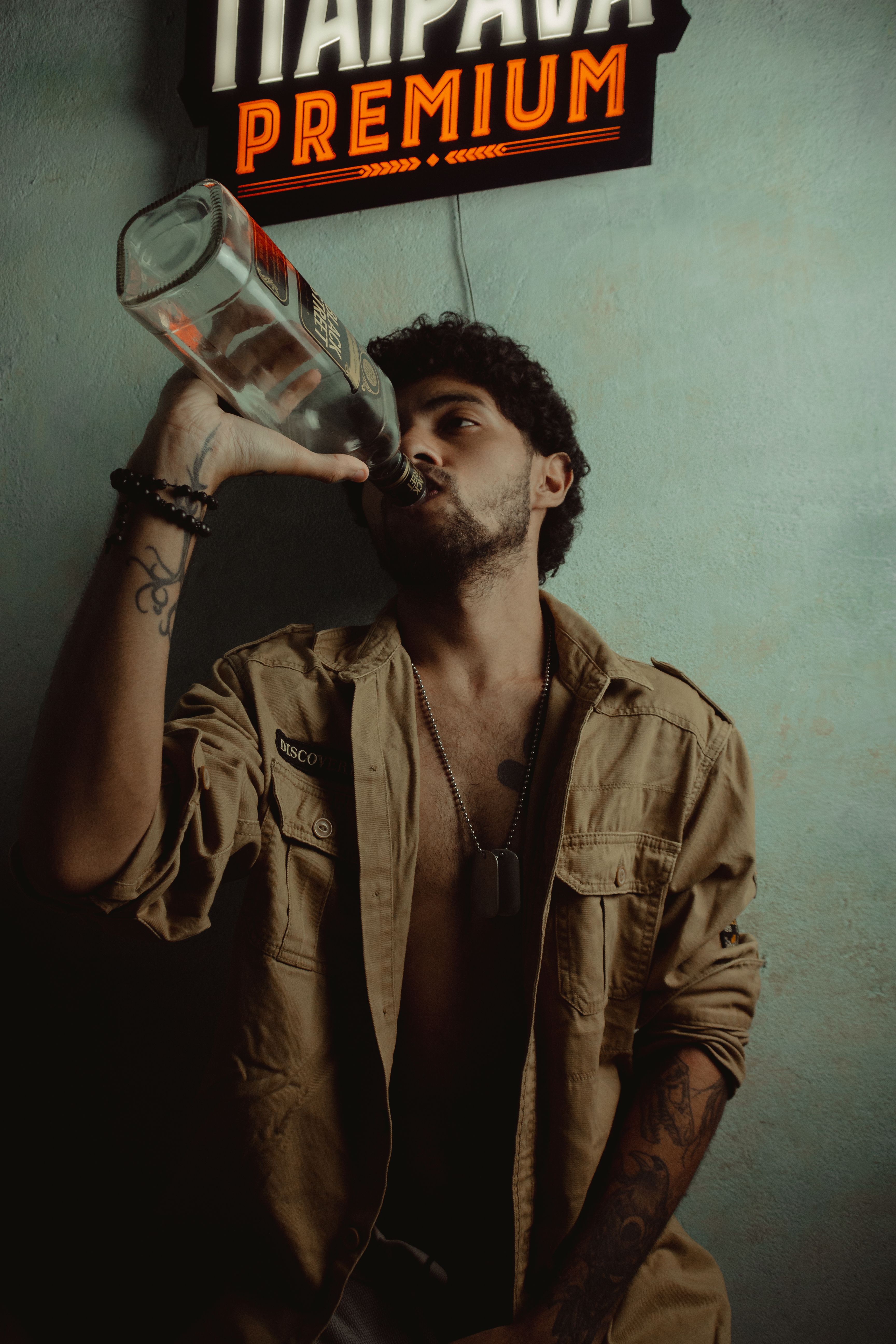 Young man in casual attire drinks from a glass bottle beneath a branded sign.