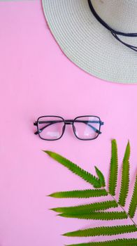 Modern eyeglasses with straw hat and fern leaf on pink background.