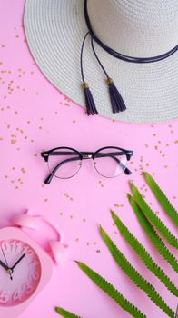 Flat lay of sunglasses, hat, and alarm clock on pink background with green leaves.