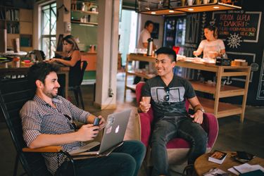 Two men having a conversation while sitting on chairs at a cofffe shop on a busy morning.