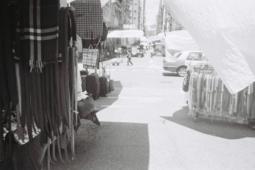 Black and white image of a bustling outdoor market in Hong Kong, showcasing textiles and street activity.