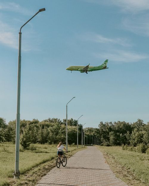 Free A woman cycles through a park as a green airplane flies overhead on a sunny day. Stock Photo