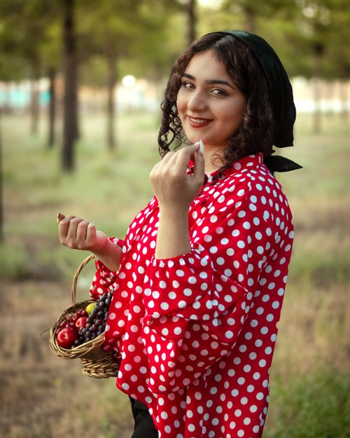 Free Smiling woman in a red polka dot blouse holding a basket of fruits outdoors in Tehran. Stock Photo
