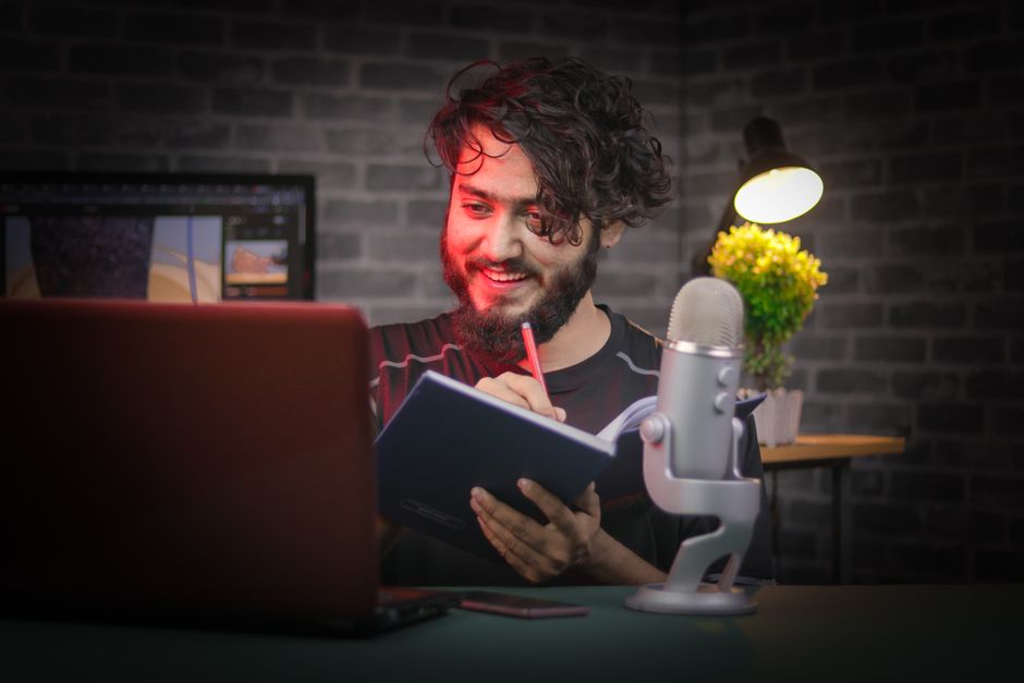 A bearded man writes notes in a cozy home office with a laptop and microphone. A bearded man writes notes in a cozy home office with a laptop and microphone.