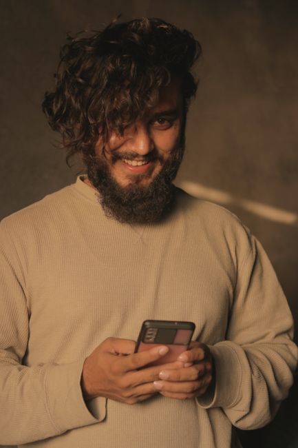 Portrait of a smiling man with curly hair using a smartphone indoors, wearing a casual sweater.
