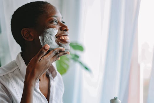 An African woman smiles while applying skincare cream to her face indoors.