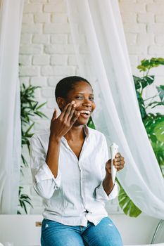 Happy woman in white blouse applying facial cream while sitting indoors, surrounded by plants.
