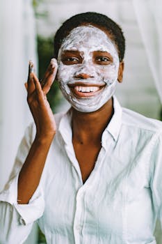 Cheerful woman with an afro hairstyle applying a face mask indoors, showcasing her skincare routine and healthy skin.