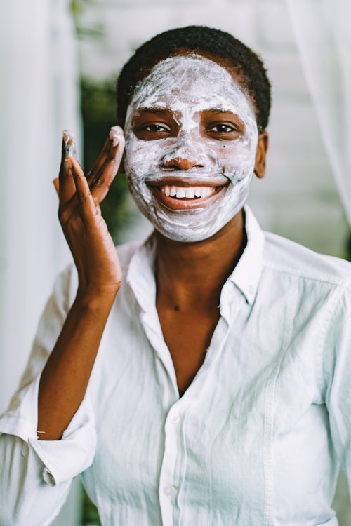 Free Cheerful woman with an afro hairstyle applying a face mask indoors, showcasing her skincare routine and healthy skin. Stock Photo