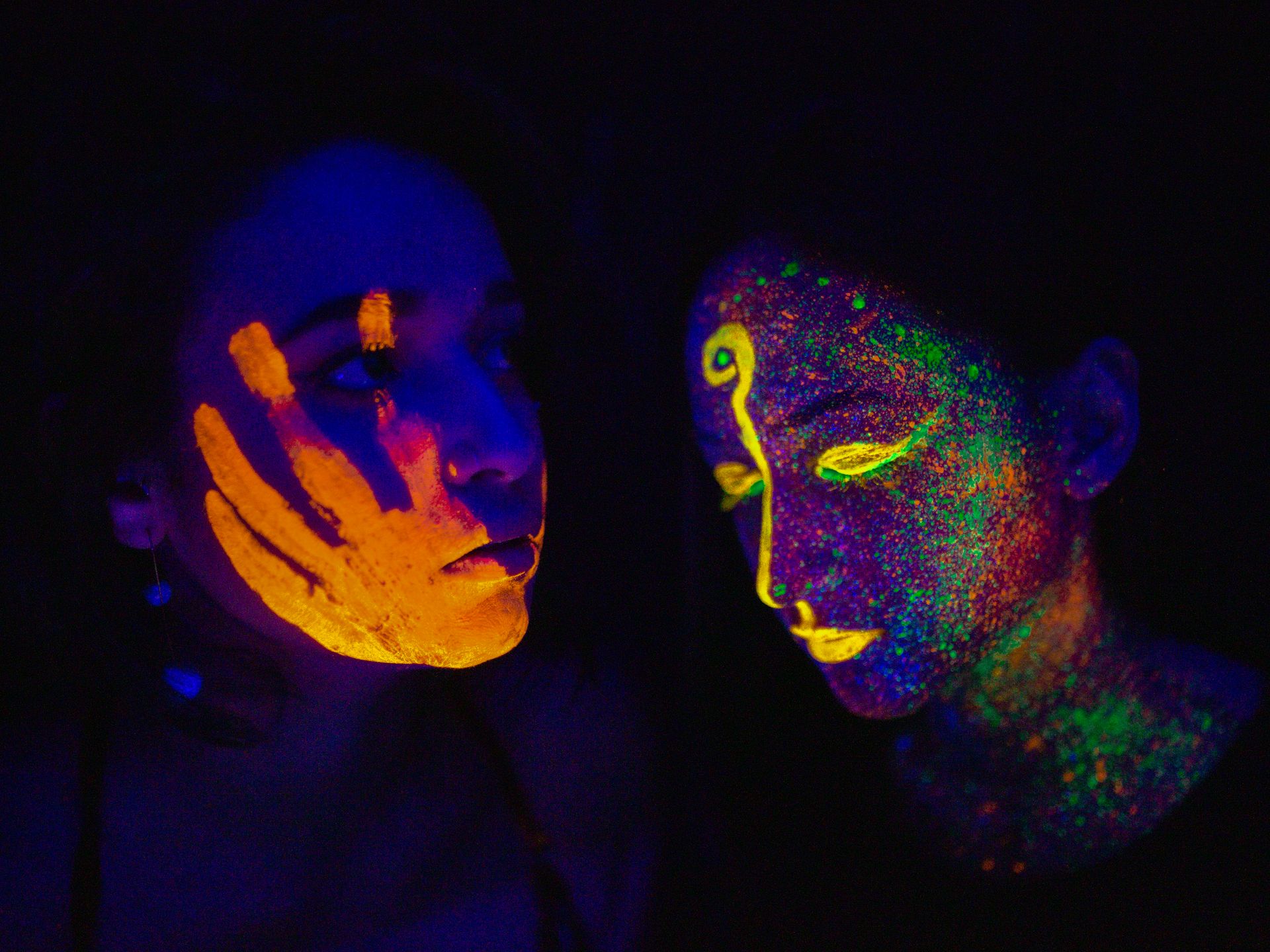 Colorful face paint glows under UV light, showcasing unique designs on two women's faces.