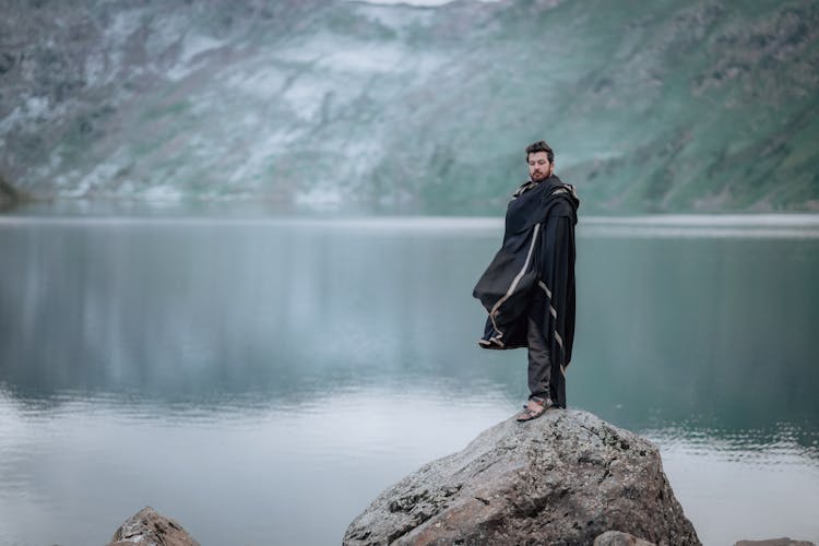 Blue Toned Landscape With Mountain Lake, And Man Wearing A Cape Standing On A Rock