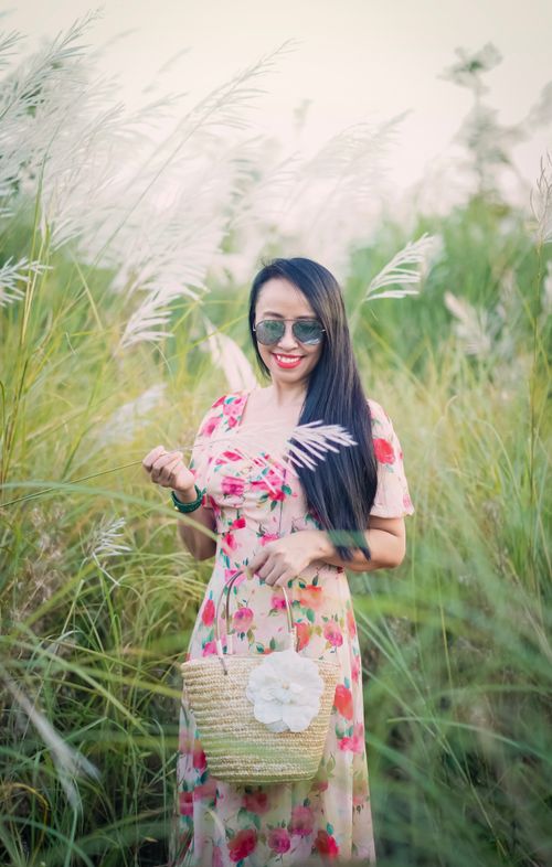Free A woman in sunglasses and floral dress poses happily in a lush green field on a sunny day. Stock Photo