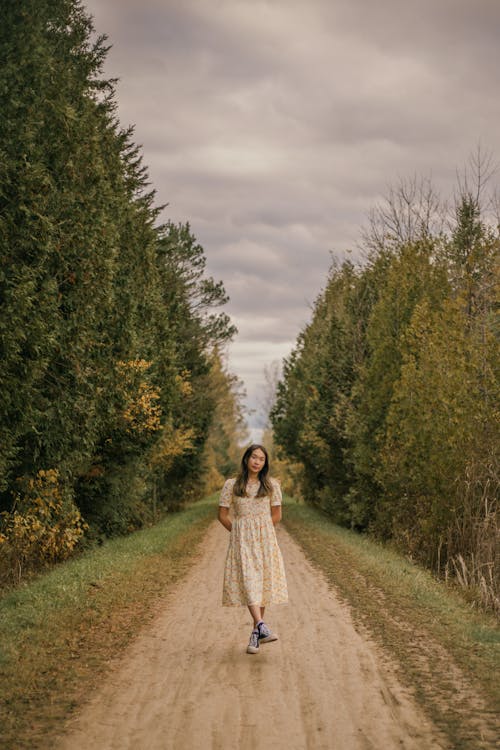 Woman in White Dress Standing on Pathway Between Green Trees Under White Clouds