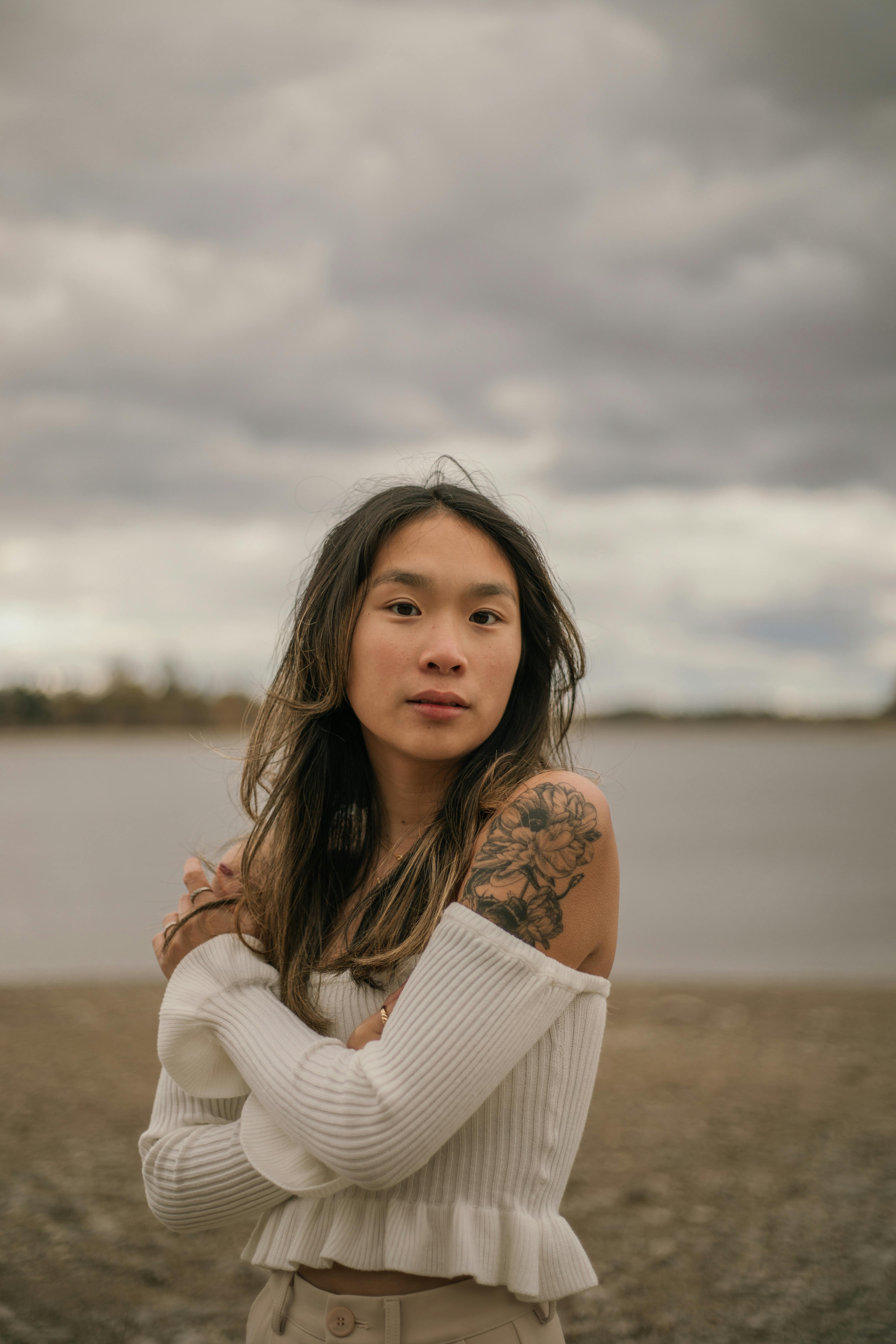 Woman with tattoos stands at the beach on an overcast day, exuding introspection and calm.