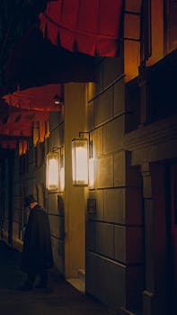 A man walks past a building with illuminated lamps at night, creating a cinematic effect.