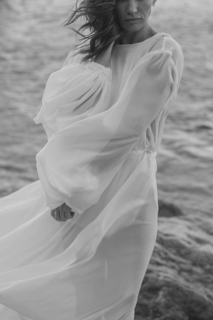 A woman in a flowing dress stands by the sea with wind-blown hair in black and white.