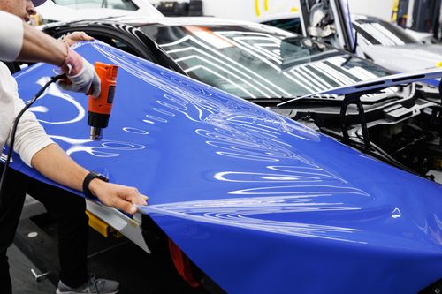 Free A technician uses a heat gun to carefully apply a blue vinyl wrap to a car in an auto workshop. Stock Photo