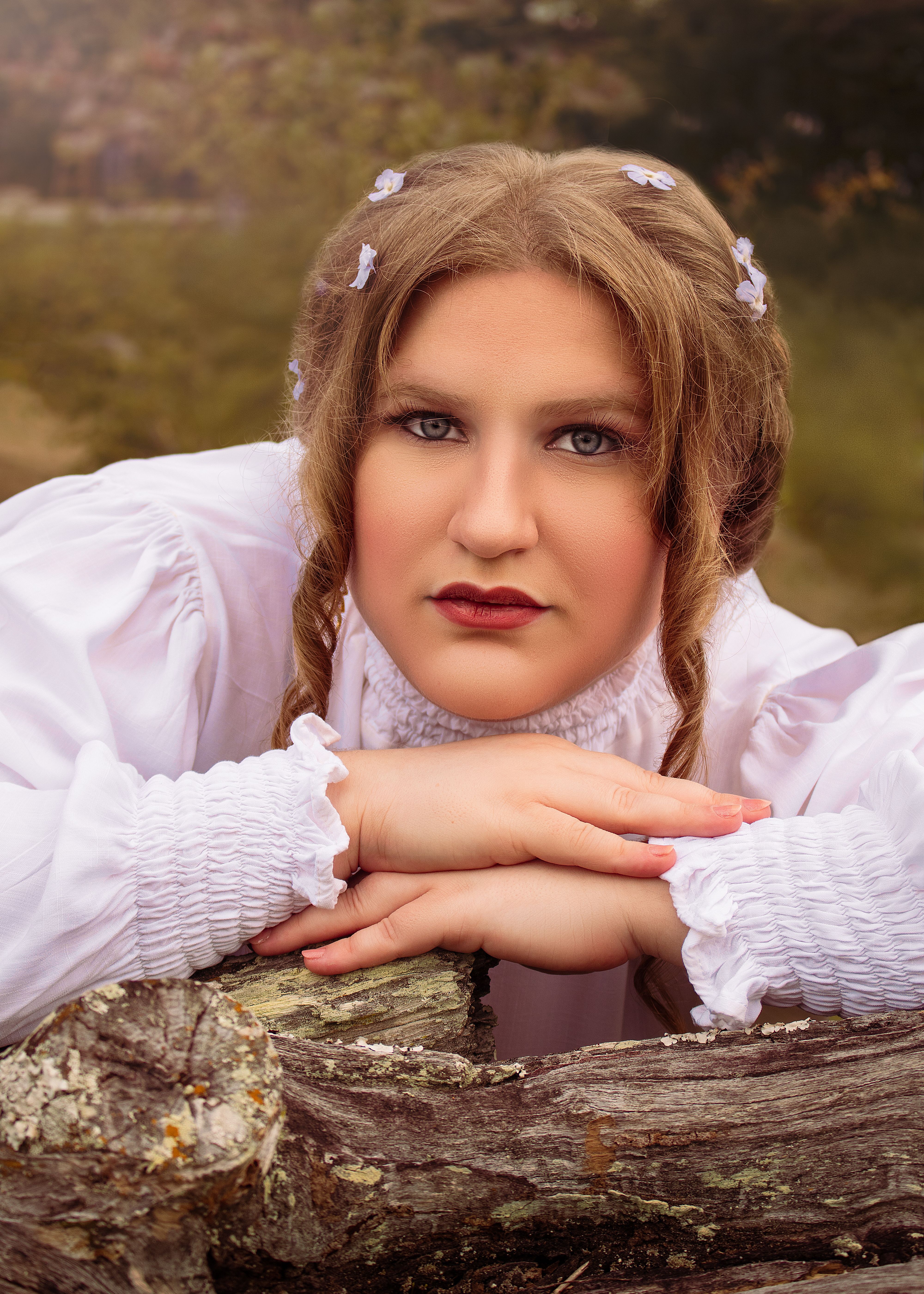 A woman with braided hair and flowers wearing a white blouse poses naturally outdoors.