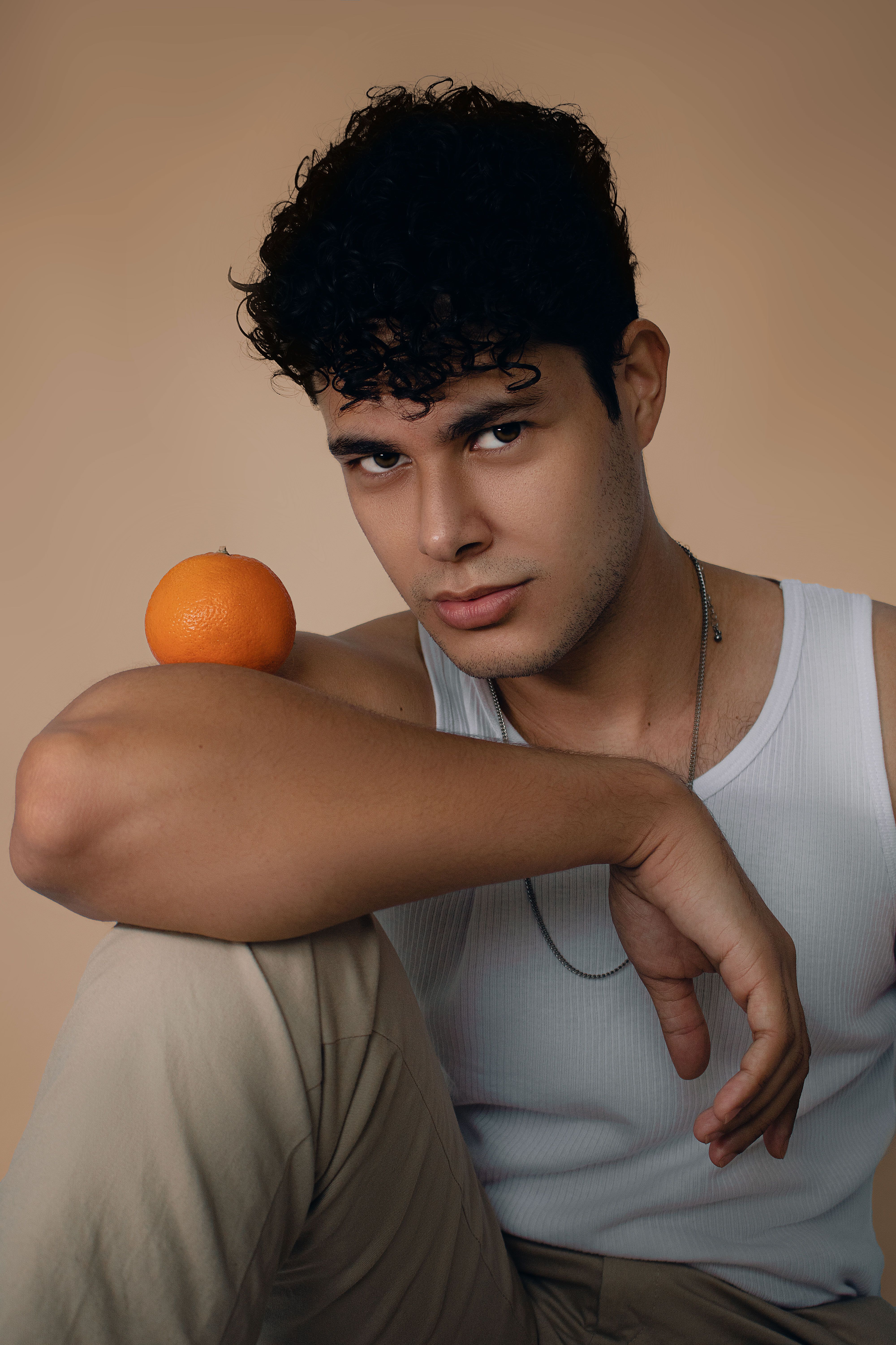 Portrait of a young man in a white tank top with an orange on his shoulder, set against a neutral background.