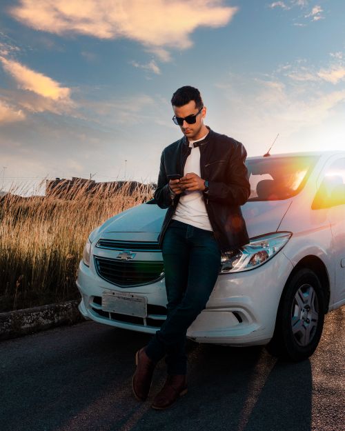 Free Man leaning on white car, using phone at sunset. Fashionable and relaxed atmosphere. Stock Photo