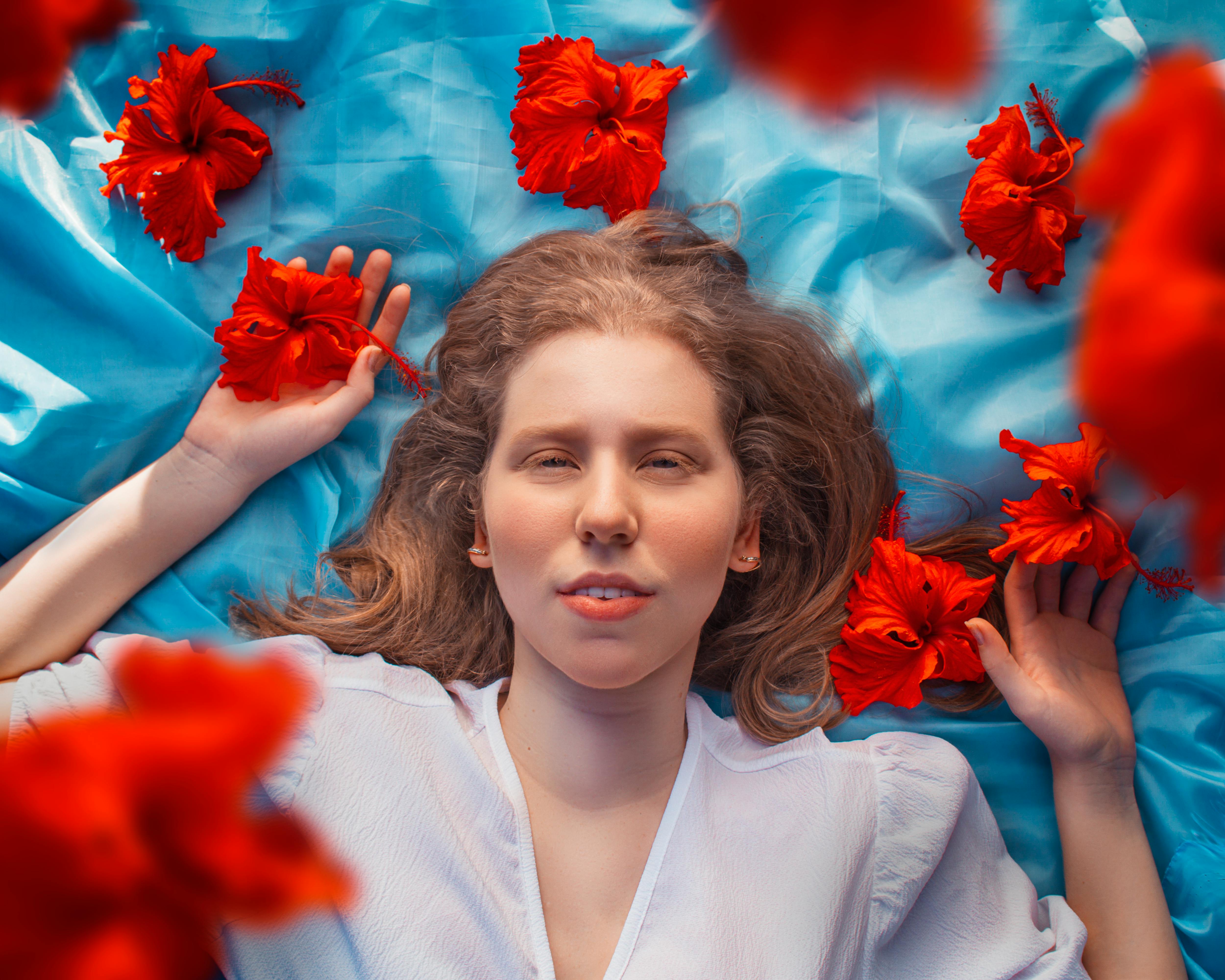 Portrait of a woman in a white dress lying on blue fabric with vibrant red hibiscus flowers.