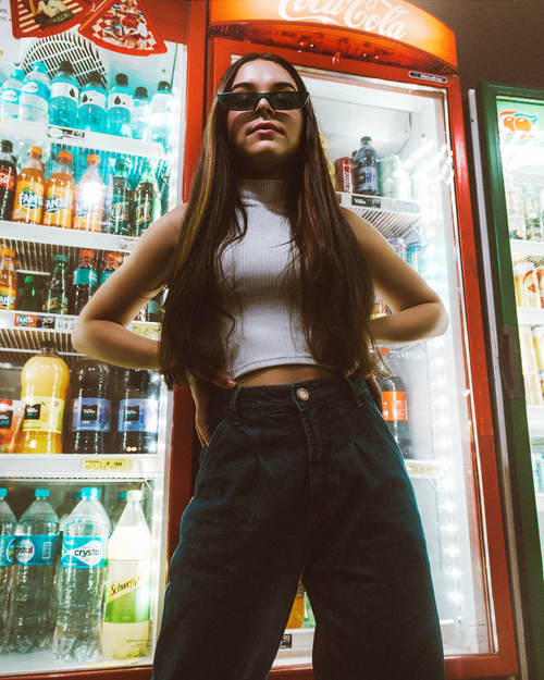 Free Fashionable young woman in sunglasses posing confidently in front of a fridge stocked with colorful beverages. Stock Photo