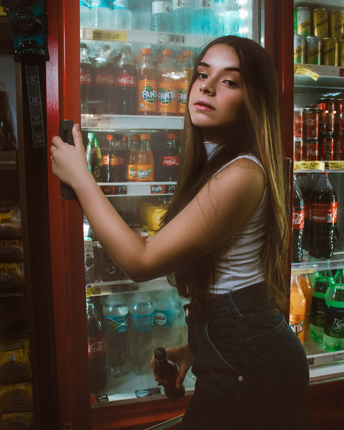 Free A young woman in casual attire poses confidently by a fridge displaying various beverages in a supermarket. Stock Photo